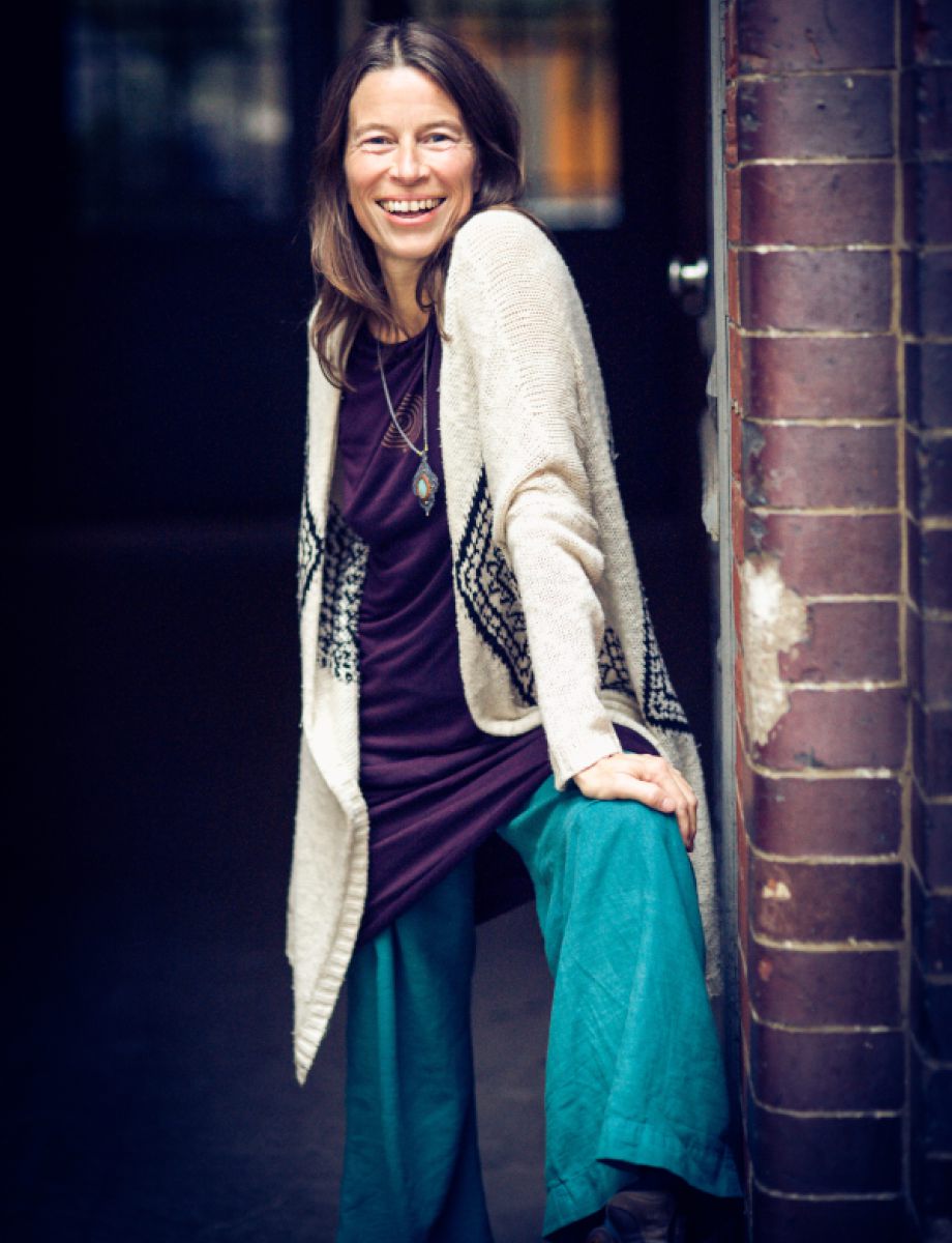 Susanne Brian steht mit blauer Hose und weißer Strickjacke vor einer Ziegelwand und lächelt in die Kamera.